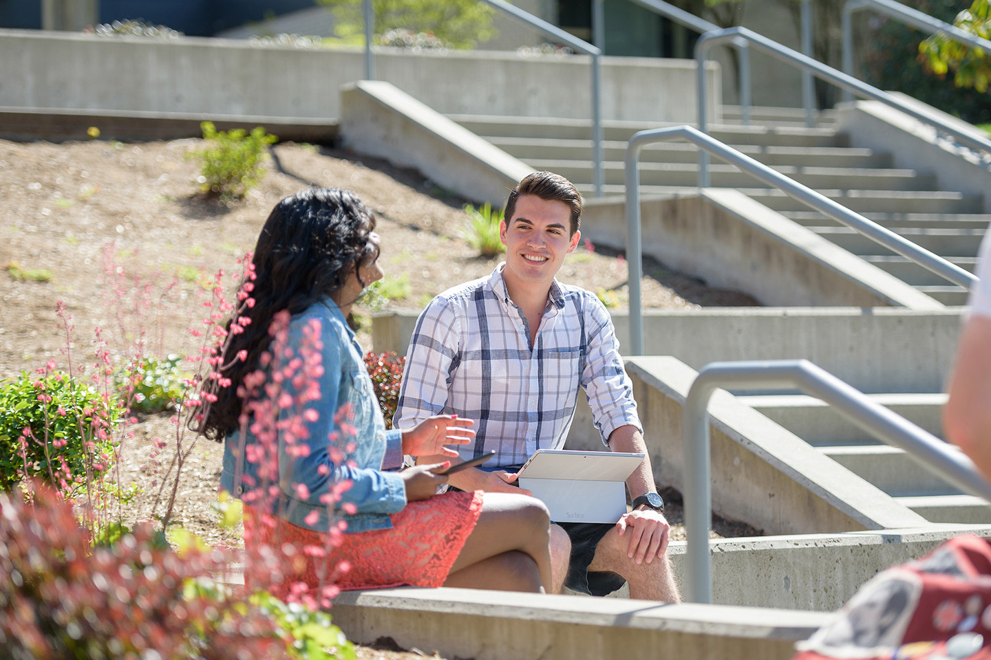 A male student holding a tablet talks with a friend outside on a bright sunny day.