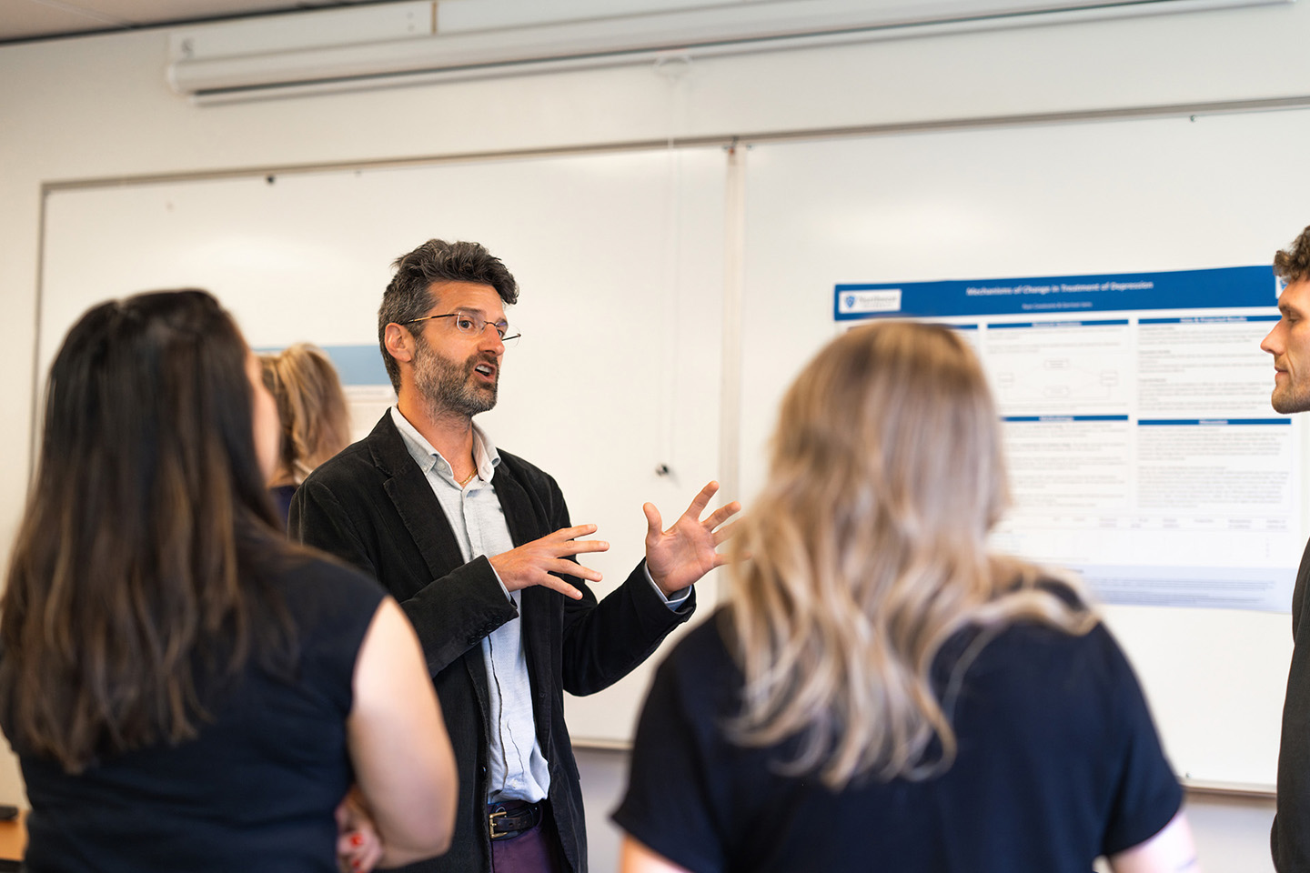 A man with glasses and a sport coat gives a presentation to fellow graduate students in a classroow.